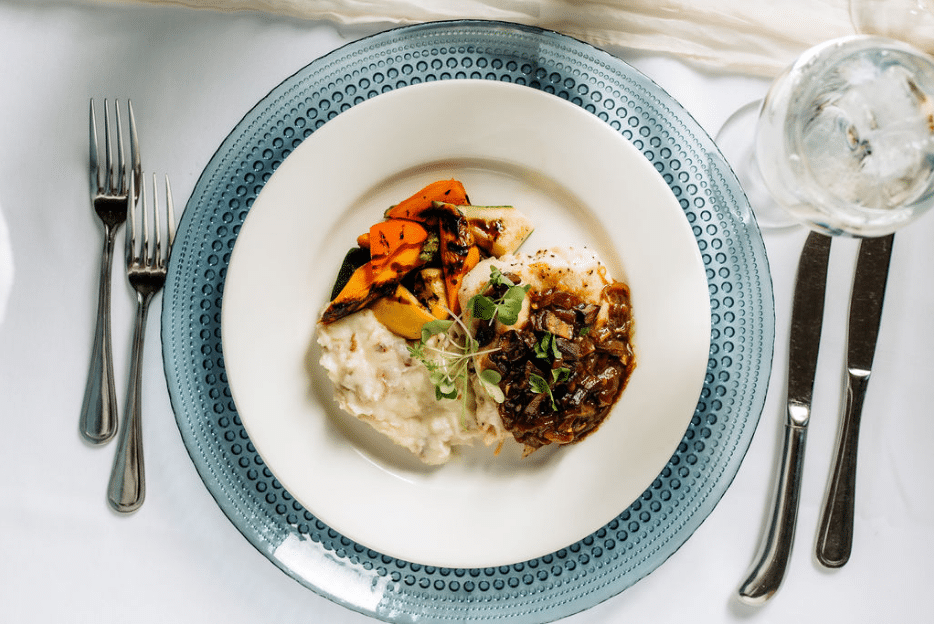 Plate of gourmet catering food, featuring mashed potatoes, roasted vegetables, and a mushroom sauce entrée on a blue charger plate.